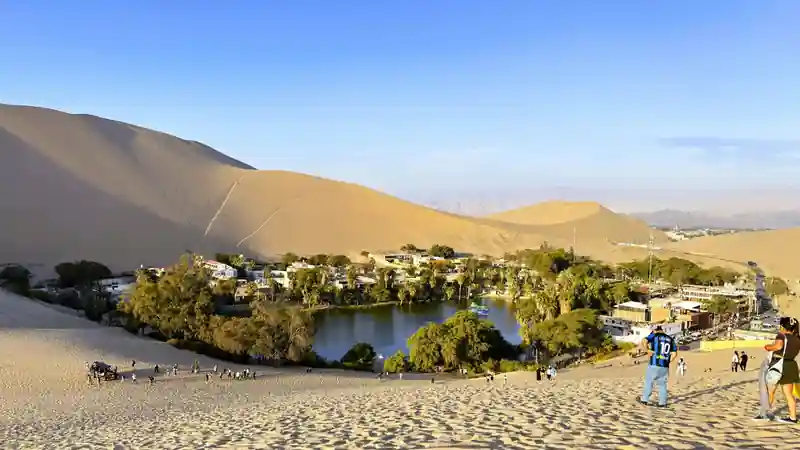 Turistas caminando sobre la arena de las dunas con vista completa al Oasis de Huacachina en Ica Peru.